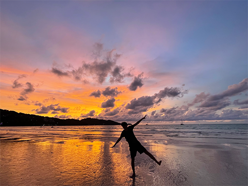 a person in shadow standing on a beach at sunset, arms wide balancing on one leg. the sky goes from blue to purple to orange to yellow